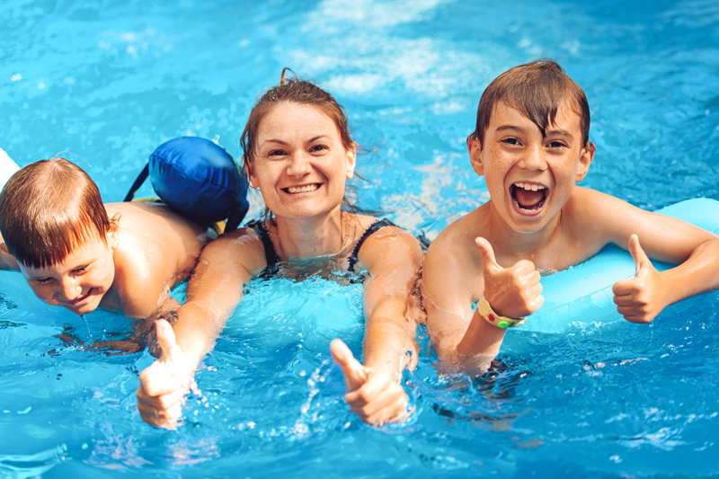 Eine lächelnde Frau mit zwei Kindern in der Therme, alle drei liegen im türkisblauen Wasser auf einer Luftmatratze und zeigen fröhlich den Daumen nach oben